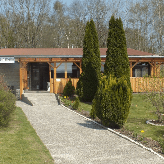 Vereinsheim mit Holzveranda, eingerahmt von Grünflächen und Sträuchern, vor einem klaren, blauen Himmel.