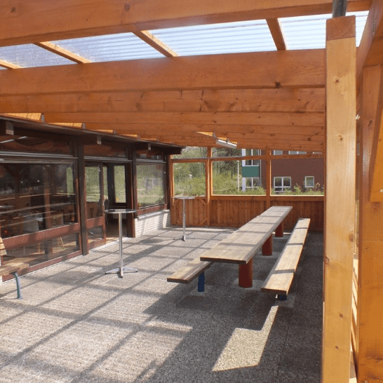 Überdachte Holzterrasse mit mehreren langen Tischen und Bänken, umgeben von Fensterfronten und Rasenflächen.