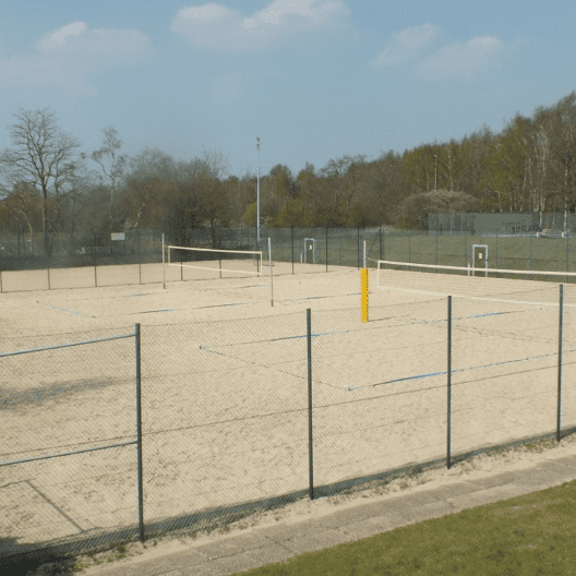 Leere Beachvolleyballfelder mit Netzen und gelben Pfosten, umgeben von einem niedrigen Zaun und Bäumen im Hintergrund.