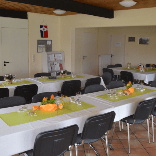 Gesellschaftsraum mit mehreren Tischen, dekoriert mit grünen Tischdecken und Blumenarrangements, helle Wände und Möbel.