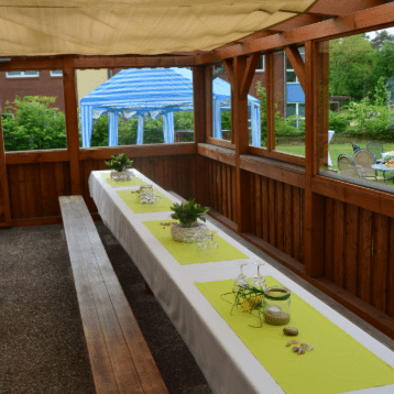 Blick ins Freie auf eine überdachte Terrasse mit langen Tischen, dekoriert mit Pflanzen und hellgrünen Tischläufern.