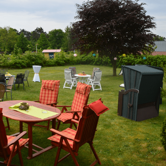 Garten mit roten Stühlen und Tisch, umgeben von Bäumen und weiteren Sitzgruppen. Zum Dekorieren hängen bunte Ballons.