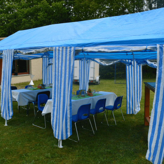 Blaue und weiße Markise über Tischen mit Stühlen auf einer Rasenfläche, im Hintergrund ein Holzgebäude.