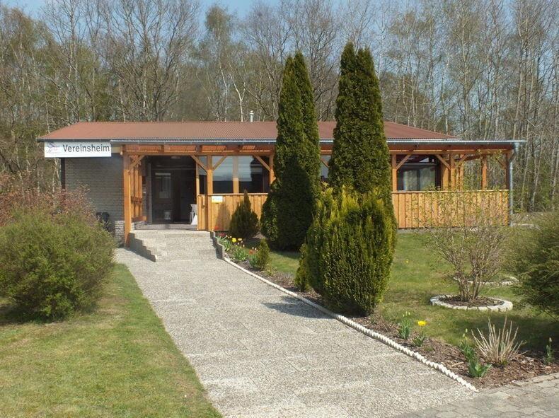 Vereinsheim Holzgebäude mit dem Schild „Vereinsheim“ und gepflegtem Gartenweg, umgeben von grünen Bäumen und Sträuchern.