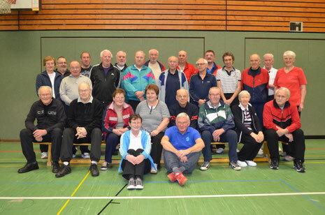 Gruppe von 24 Personen in Sportkleidung, versammelt auf einer gr&uuml;nen Sportfl&auml;che in einer Turnhalle.