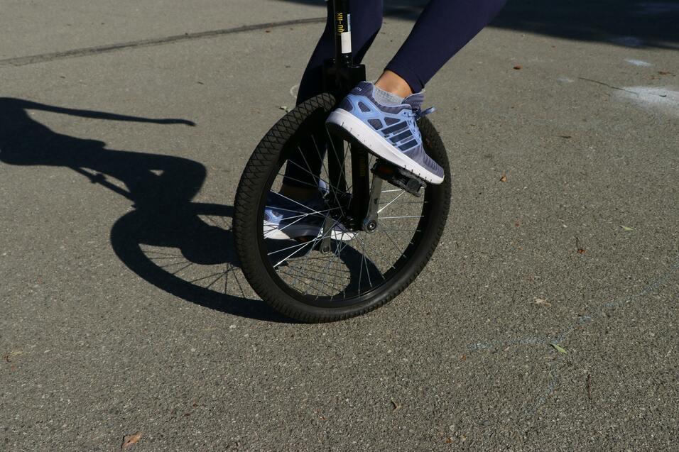 Nahaufnahme eines Fußes in einem sportlichen Schuh auf einem Einrad, das auf einer asphaltierten Fläche balanciert.