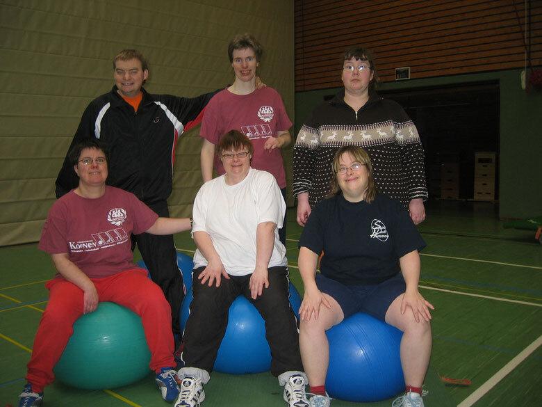 Sechs Personen sitzen auf Gymnastikbällen in einer Sporthalle, lächelnd und in sportlicher Kleidung.