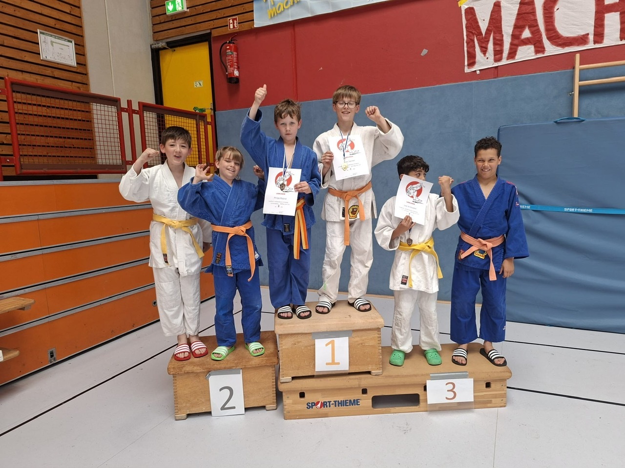 Sechs Kinder in Judogi stehen auf einem Siegertreppchen und halten Urkunden, l&auml;chelnd und jubelnd.