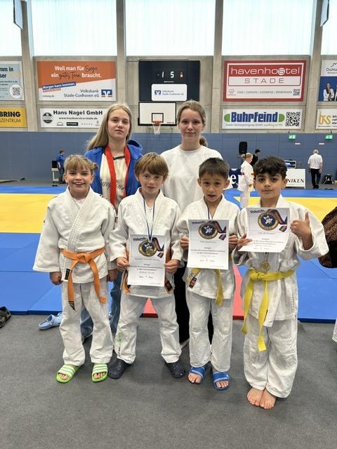 Gruppe von fünf Judokas in weißen Anzügen mit Urkunden und Medaillen auf einer Judomatte in einer Halle.