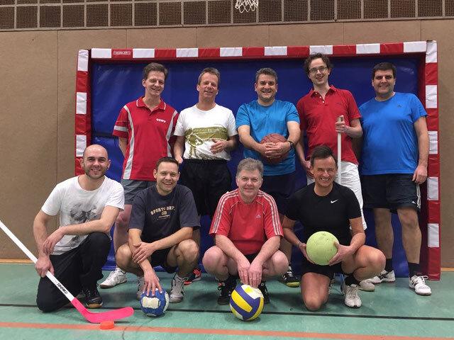 Gruppe von neun sportlichen Männern mit verschiedenen Bällen in einer Sporthalle vor einem Tor.