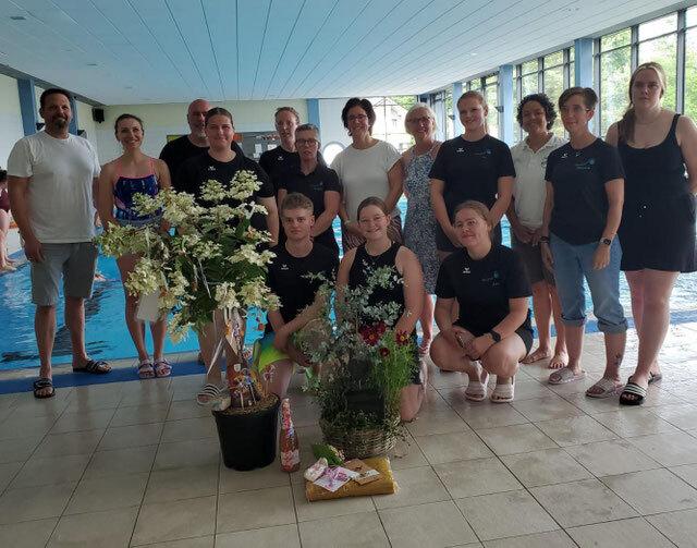 Gruppe von Schwimmern und Trainern in Schwimmbad, umgeben von Blumenarrangements und Geschenken.