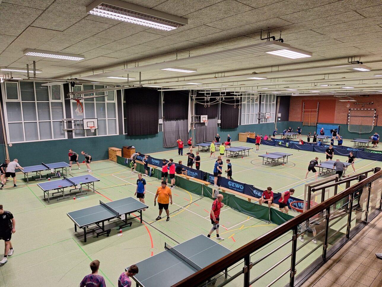 Volles Tischtenniszentrum mit mehreren Tischen und Spielern in Aktion, von einer erhöhten Perspektive aufgenommen.