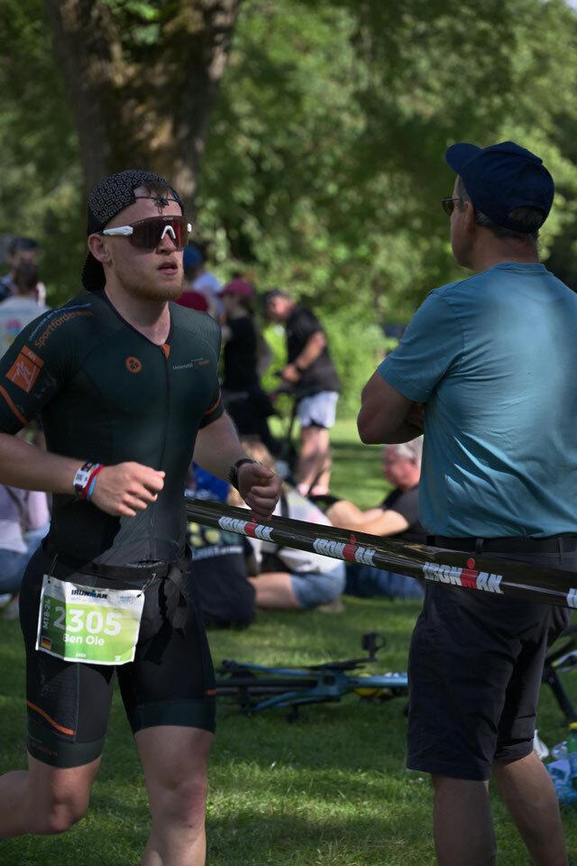 Sportler in Triathlonbekleidung läuft vorbei, während ein Zuschauer im Hintergrund zu ihm spricht. Grüne Wiese, Wettkampfatmosphäre.