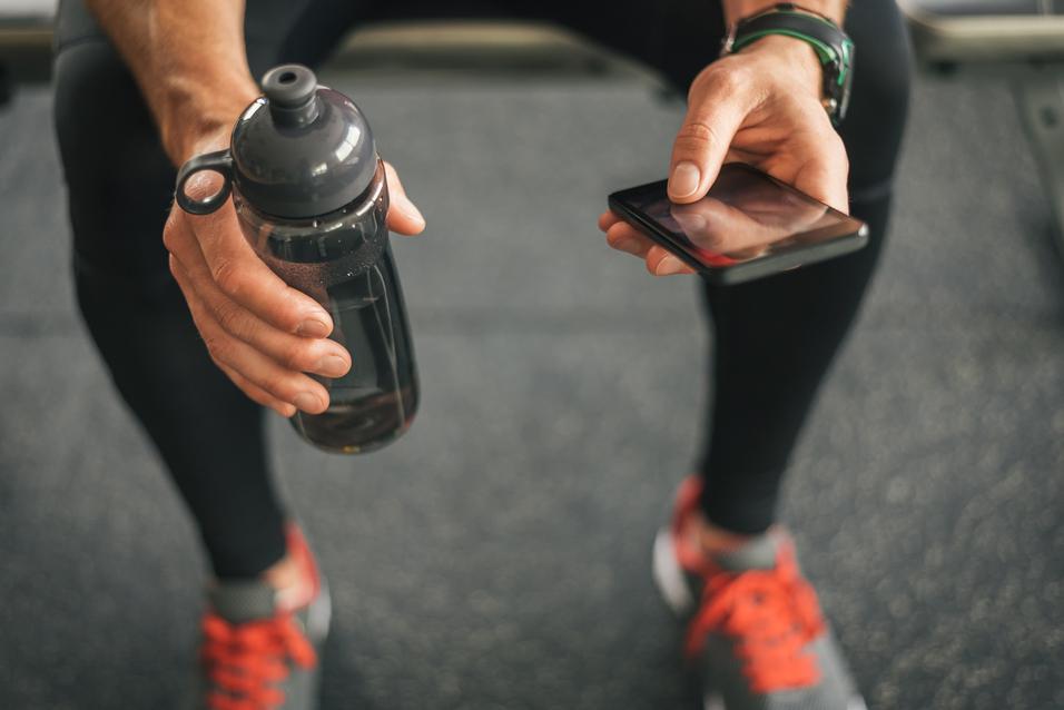 Nahaufnahme einer sportlichen Hand, die eine Trinkflasche hält, während die andere ein Smartphone berührt.