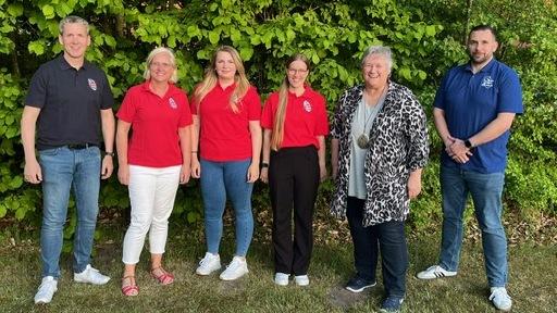 Sechs Personen stehen vor einem grünen Hintergrund aus Laub, alle tragen unterschiedliche Kleidungsstücke in rot und blau.