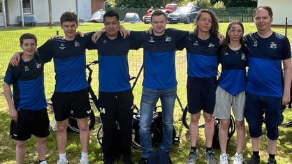 Gruppe von sieben Personen in blauen Trikots steht im Freien, umgeben von Fahrrädern und grüner Landschaft.