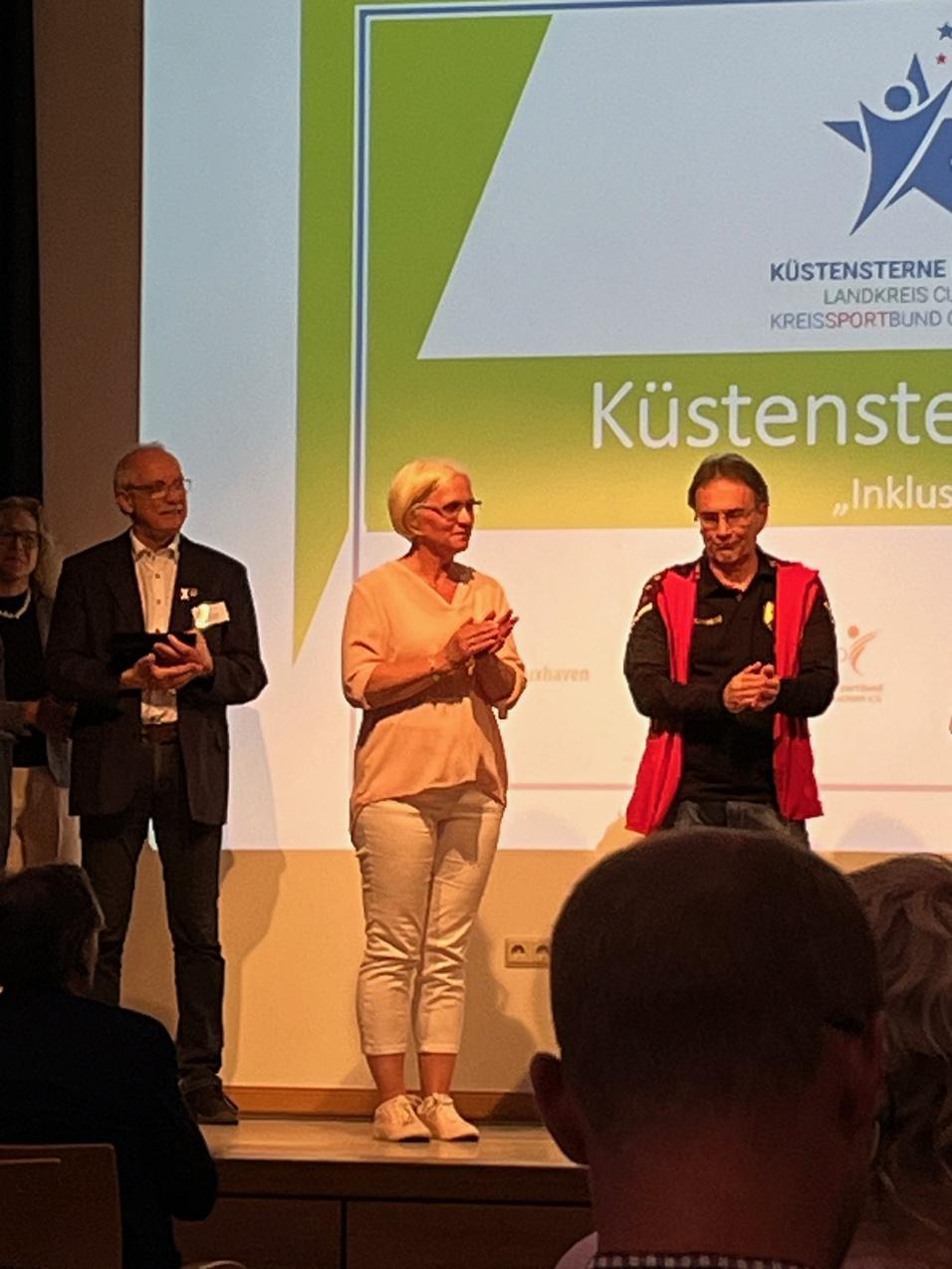 Eine Gruppe von Personen steht auf einer Bühne, während im Hintergrund ein Präsentationsbildschirm sichtbar ist.