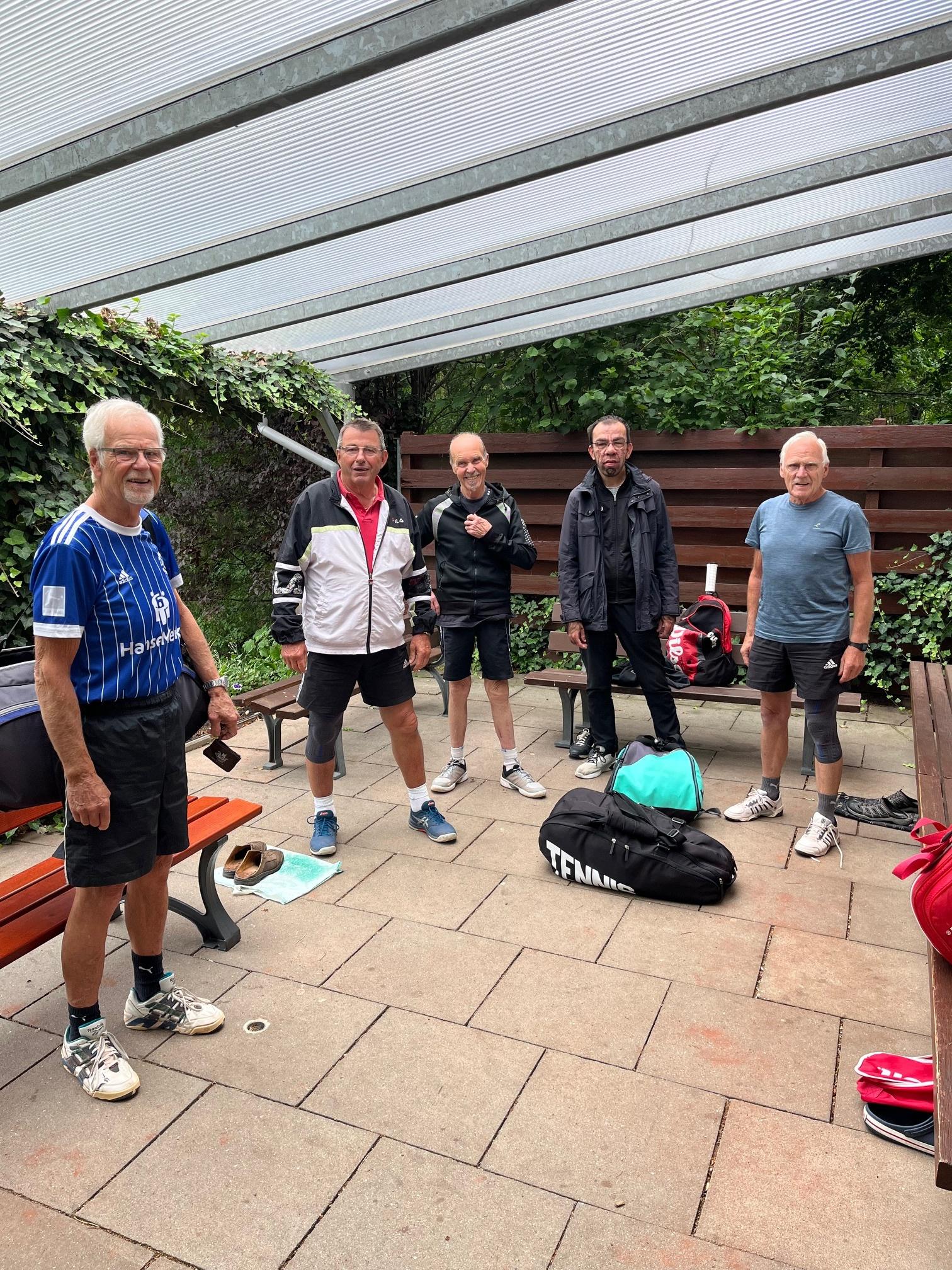 Fünf ältere Männer stehen in einer überdachten Außenfläche, umgeben von Pflanzen und Sporttaschen.