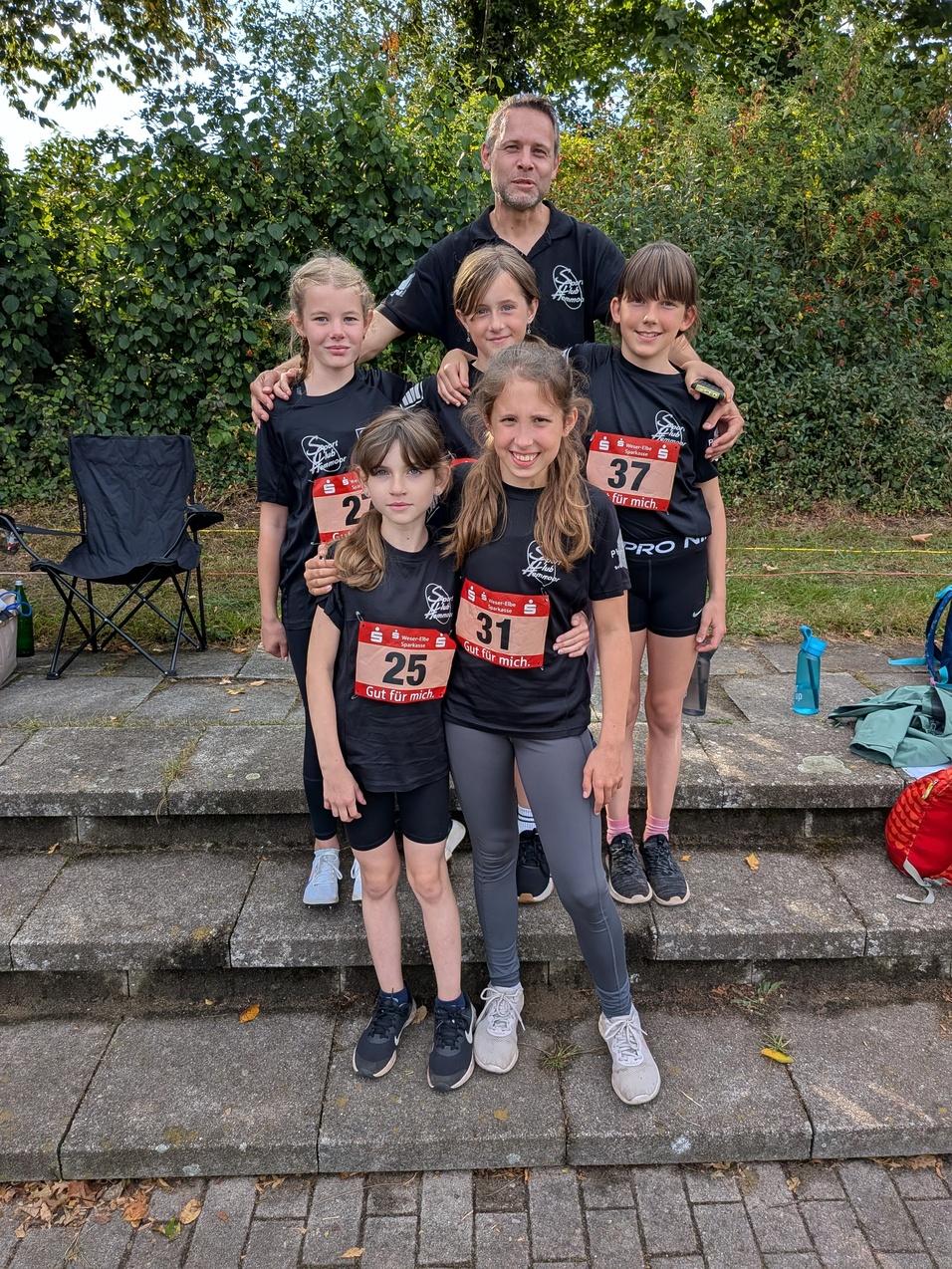 Sechs Kinder und ein Erwachsener in Sportkleidung, stehen auf einer Treppe nach einem Wettkampf. Alle tragen Startnummern.
