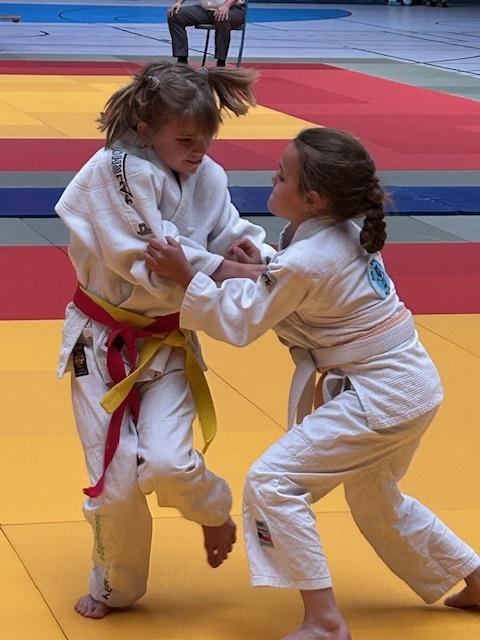 Zwei Kinder in Judoanzügen kämpfen auf einer farbigen Judomatte. Eines hat einen roten, das andere einen blauen Gürtel.