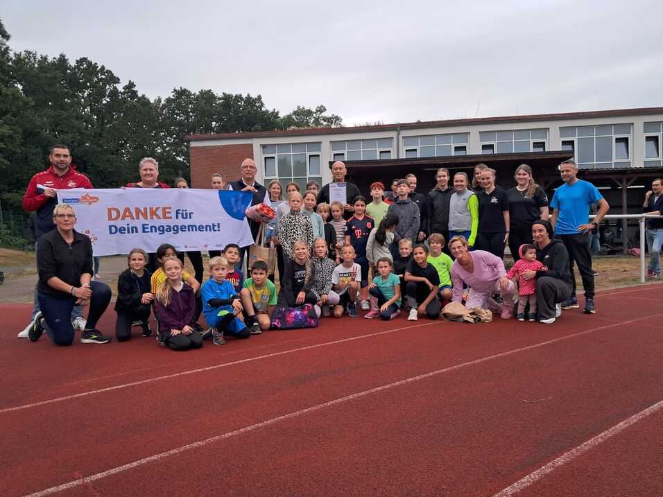 Gruppe von Menschen, darunter Kinder und Erwachsene, posieren auf einer Laufbahn mit einem Banner „DANKE für Dein Engagement!“.