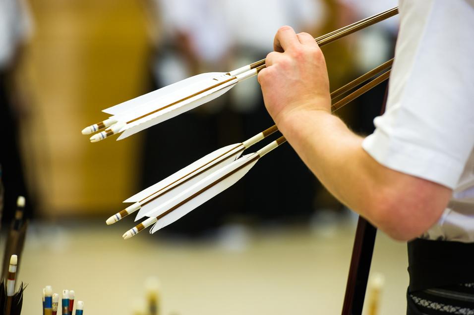 Nahaufnahme einer Hand, die mehrere Pfeile mit weißen Federn in einer Sportumgebung hält.
