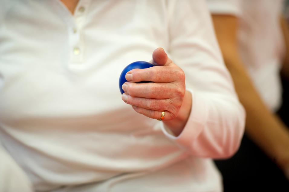 Nahaufnahme einer Hand, die einen blauen Ball hält, während die Person in einem weißen Oberteil sitzt.